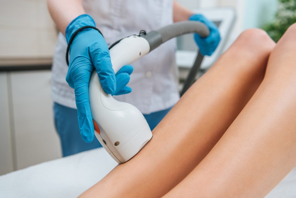 Partial view of cosmetologist in rubber gloves doing laser hair removal procedure on legs